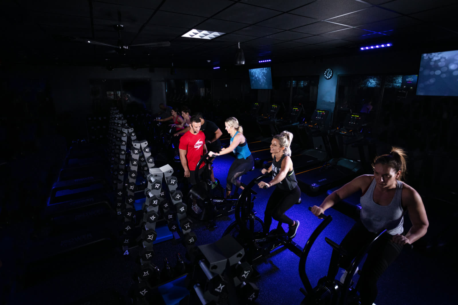 Members riding bikes and training on the dimly lit Bria Method studio floor under blue lighting